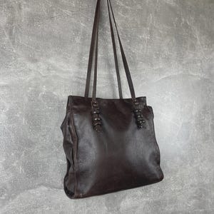 Könnte beinhalten: Eine dunkelbraune Ledertasche mit langen Trägern, die an einer grauen Wand hängt. Die Tasche hat eine rechteckige Form mit dekorativen Details oben und ein leicht abgenutztes Aussehen.