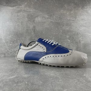 May include: A two-tone golf shoe in white and blue leather. The shoe features a wingtip design with decorative perforations and white laces. The sole has small rubber cleats for traction. The shoe is designed for the golf course.
