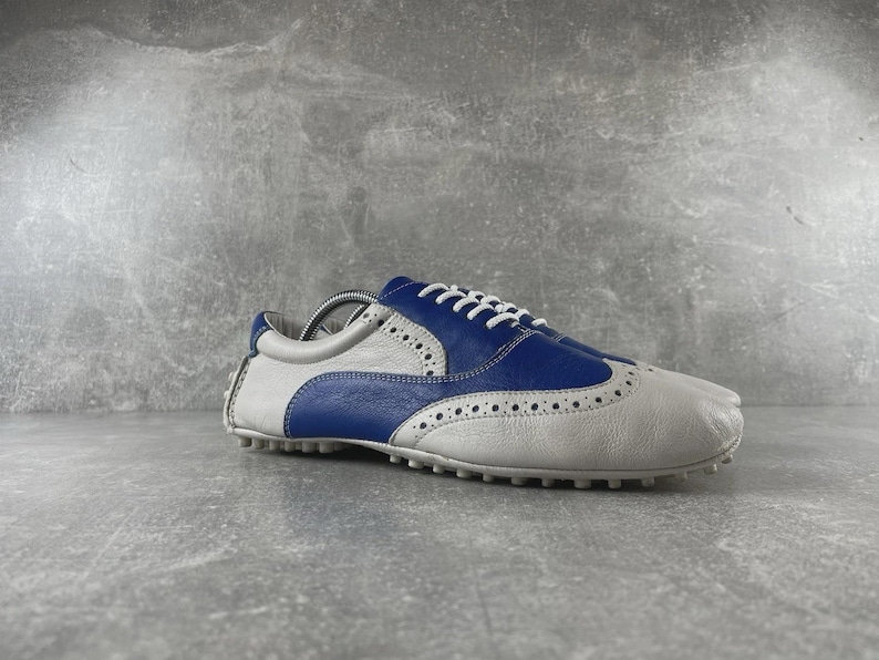 May include: A two-tone golf shoe in white and blue leather. The shoe features a wingtip design with decorative perforations and white laces. The sole has small rubber cleats for traction. The shoe is designed for the golf course.