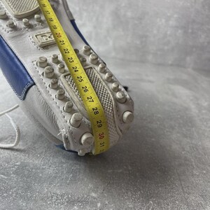 May include: White leather driving shoe with blue detailing. The sole has small, rounded rubber studs. A yellow measuring tape shows the shoe's length. The shoe has a white lace and the brand name is visible.