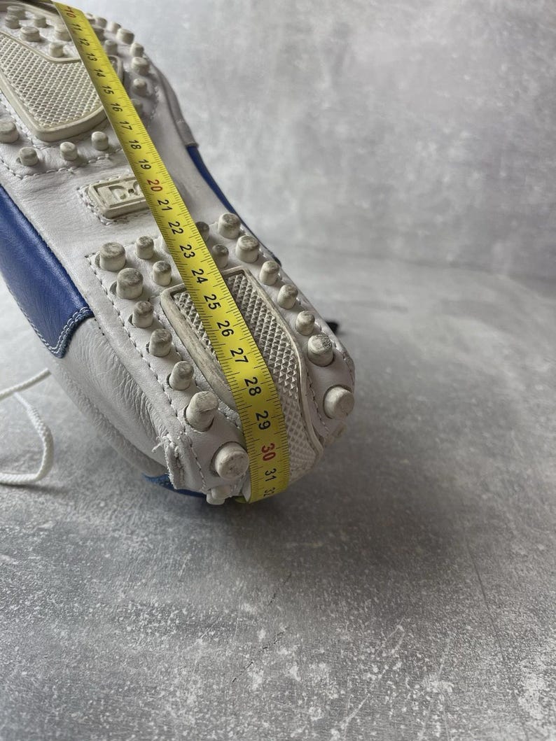 May include: White leather driving shoe with blue detailing. The sole has small, rounded rubber studs. A yellow measuring tape shows the shoe's length. The shoe has a white lace and the brand name is visible.