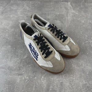 May include: A pair of white and beige sneakers featuring black laces and the Von Dutch logo embroidered in blue on the side. The shoes have a low-top silhouette with suede overlays on the toe and heel, a padded collar, and a brown rubber sole.