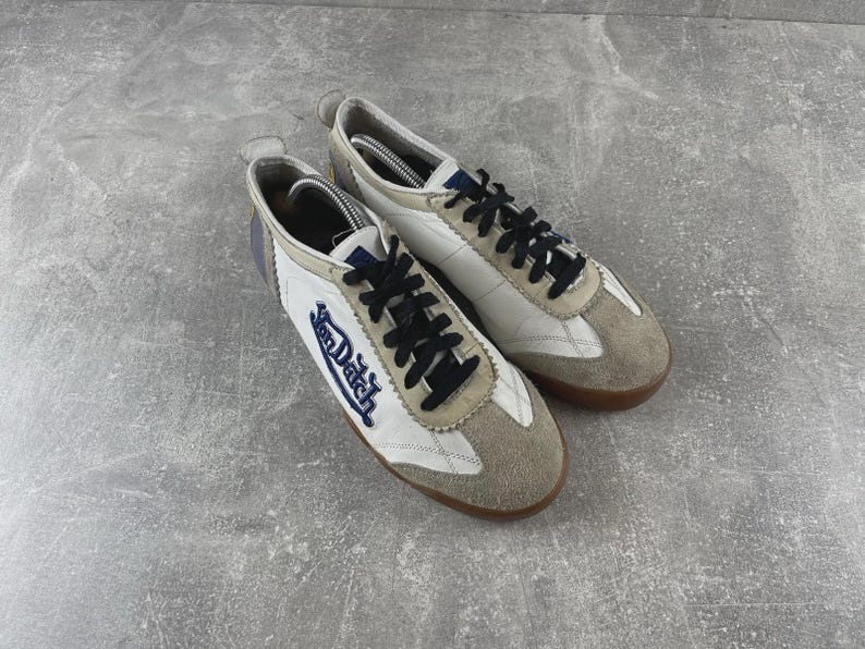 May include: A pair of white and beige sneakers featuring black laces and the Von Dutch logo embroidered in blue on the side. The shoes have a low-top silhouette with suede overlays on the toe and heel, a padded collar, and a brown rubber sole.