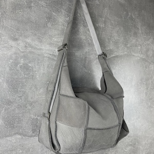 Pode incluir: Uma bolsa de ombro cinza com patchwork, uma alça longa e um zíper prateado. A bolsa é feita de couro e tem um design geométrico único. A bolsa está pendurada em uma parede cinza.