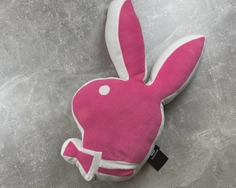 May include: A plush cushion shaped like a rabbit head silhouette in bright pink with white trim along the ears and base. The design features a circular white eye detail and a small black fabric tag attached to the side with white text.