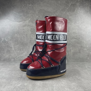 Peut inclure: Une paire de Moon Boots rouges et bleu marine avec le nom de la marque en lettres blanches sur une bande blanche. Les bottes ont des lacets noirs et une semelle beige. Les bottes sont conçues pour le froid.