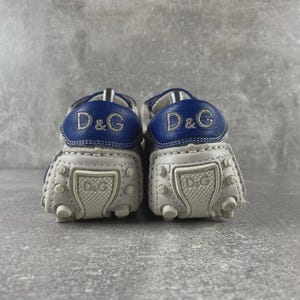May include: A pair of white and blue leather baby shoes. The shoes have a rounded toe design with small rubber studs on the sole. The heel features a blue leather patch with the embroidered letters "D&G".