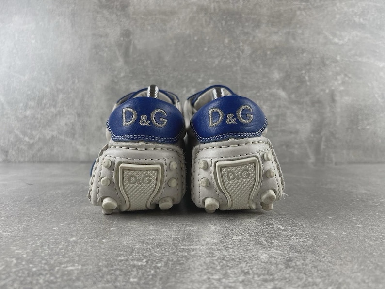May include: A pair of white and blue leather baby shoes. The shoes have a rounded toe design with small rubber studs on the sole. The heel features a blue leather patch with the embroidered letters "D&G".