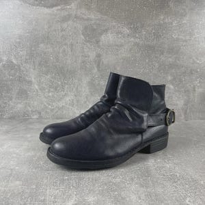 May include: A pair of dark blue ankle boots. The boots have a rounded toe, a low heel, and a strap with a buckle on the side. The leather is slightly wrinkled, giving the boots a worn look.