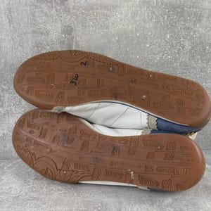May include: A pair of white sneakers with dark blue heel accents and brown rubber outsoles. The outsoles feature a repeating embossed pattern of small rectangular logos and text. The white upper material is stitched with a subtle diagonal pattern.