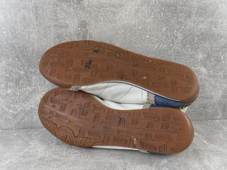 May include: A pair of white sneakers with dark blue heel accents and brown rubber outsoles. The outsoles feature a repeating embossed pattern of small rectangular logos and text. The white upper material is stitched with a subtle diagonal pattern.