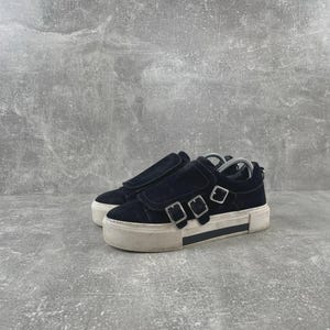 May include: A pair of black suede sneakers featuring a thick white rubber platform sole and three silver-toned metal buckles across the top. The footwear has a rounded toe design and a low-top silhouette with a padded collar and a visible shoe tree.