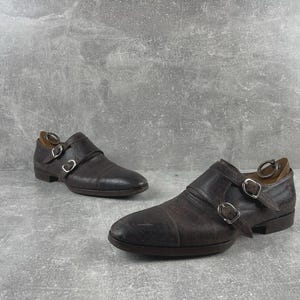 May include: A pair of brown leather double monk strap shoes. The shoes have two silver buckles and a rounded toe. The leather has a textured appearance. The shoes are set against a gray background.