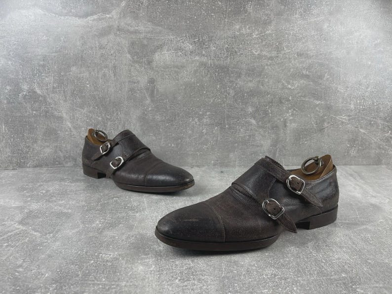 May include: A pair of brown leather double monk strap shoes. The shoes have two silver buckles and a rounded toe. The leather has a textured appearance. The shoes are set against a gray background.
