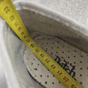 May include: A white shoe interior featuring a perforated insole with the partial text "tch" printed in black. A yellow measuring tape is placed inside, showing a length measurement extending from 18 to 33 centimeters along the shoe's inner base.