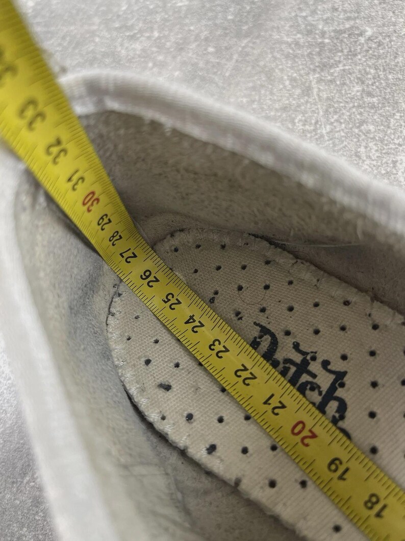May include: A white shoe interior featuring a perforated insole with the partial text "tch" printed in black. A yellow measuring tape is placed inside, showing a length measurement extending from 18 to 33 centimeters along the shoe's inner base.