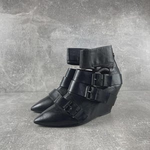 May include: Black leather ankle boots with multiple straps and buckles. The boots have a pointed toe and a wedge heel. The design includes a high ankle cuff and a zipper at the back.