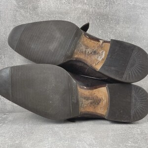 May include: A pair of worn, dark brown leather dress shoes, soles facing up. The soles are black with a textured pattern for grip. The heels and inner soles show signs of wear. The shoes are likely vintage or pre-owned.