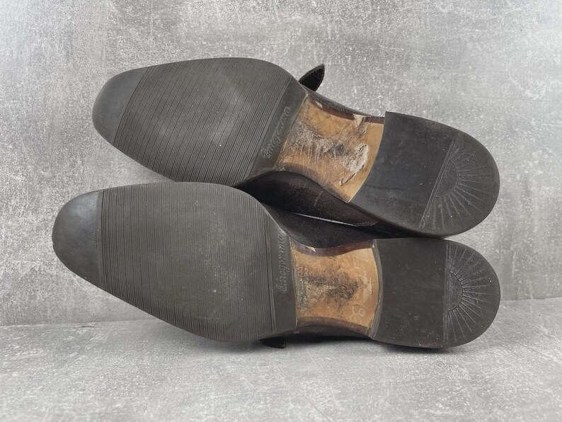 May include: A pair of worn, dark brown leather dress shoes, soles facing up. The soles are black with a textured pattern for grip. The heels and inner soles show signs of wear. The shoes are likely vintage or pre-owned.