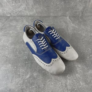 May include: Pair of two-tone leather shoes. The shoes are light grey with blue detailing on the toe and heel. White laces and decorative perforations add detail. The shoes are designed with a classic wingtip style.