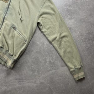 May include: A sage green zip-up hoodie with a drawstring hood and a front pocket. The hoodie has a ribbed hem and cuffs with a striped design. A tag with the text "Von Dutch" is attached to the zipper.