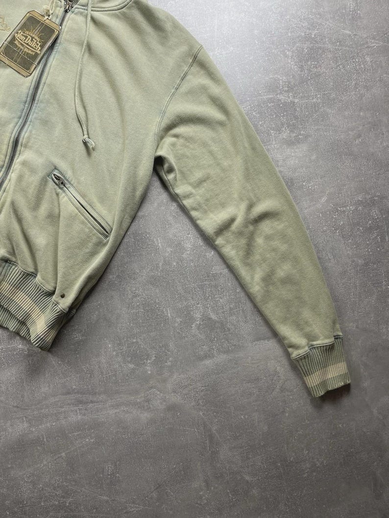 May include: A sage green zip-up hoodie with a drawstring hood and a front pocket. The hoodie has a ribbed hem and cuffs with a striped design. A tag with the text "Von Dutch" is attached to the zipper.