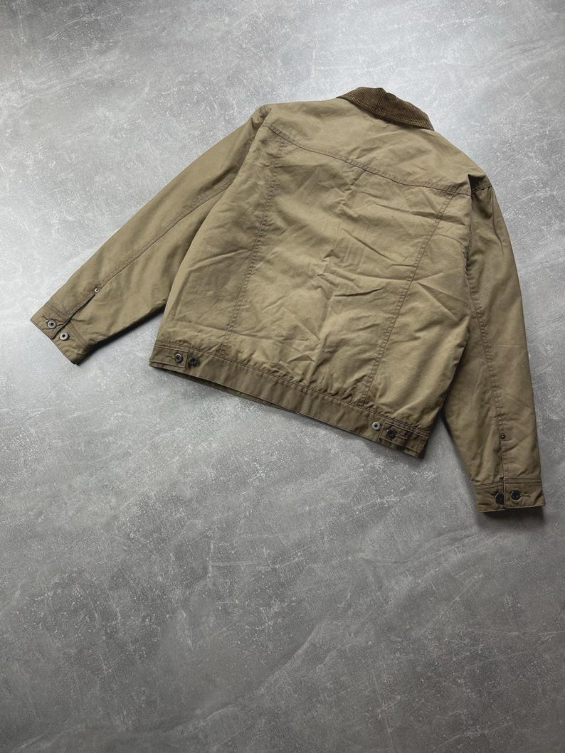 Puede incluir: Una chaqueta de color tostado con cuello de pana marr&oacute;n oscuro, mangas largas con pu&ntilde;os abotonados y dobladillo con botones. La parte posterior de la chaqueta muestra detalles de costuras horizontales y costuras verticales en el cuerpo.