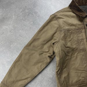 Puede incluir: Una chaqueta utilitaria de color tostado con cuello de pana marr&oacute;n oscuro, mangas largas y un bolsillo grande de parche en la parte delantera. La chaqueta est&aacute; confeccionada en un tejido mate resistente y cuenta con cierre de botones y costuras.