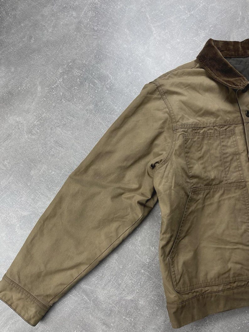 Puede incluir: Una chaqueta utilitaria de color tostado con cuello de pana marr&oacute;n oscuro, mangas largas y un bolsillo grande de parche en la parte delantera. La chaqueta est&aacute; confeccionada en un tejido mate resistente y cuenta con cierre de botones y costuras.