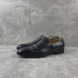 Puede incluir: Un par de mocasines de cuero negro con punta cuadrada, tacón bajo y acabado liso. Los zapatos tienen un forro interior de color beige e incluyen hormas de metal plateado insertadas en el interior para mantener su forma original.