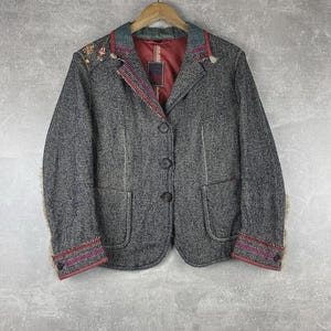 May include: A grey tweed blazer with a textured pattern, featuring a red lining and decorative stitching. The blazer has a notched collar, two front pockets, and button closures. The cuffs and collar are accented with colourful trim and embroidery.