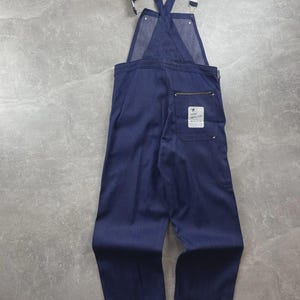 May include: Dark blue denim overalls featuring adjustable shoulder straps with metal buckles, a high back, and a single rear pocket with a white rectangular patch displaying the text QUALITY. The garment has a straight-leg cut and visible stitching.