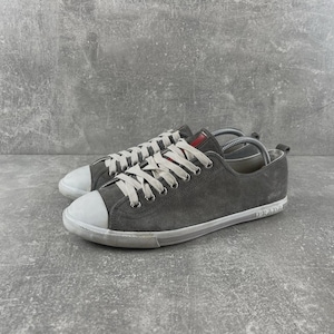 May include: A pair of low-top sneakers in dark gray suede with white rubber toe caps and white rubber soles. The shoes feature white laces, silver-tone metal eyelets, and a small red rectangular tag on the tongue. The word Prada is embossed on the heel.