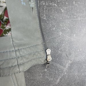 Peut inclure: Gros plan sur une veste en cuir gris clair avec une fermeture &eacute;clair argent&eacute;e. La veste pr&eacute;sente des d&eacute;tails floraux peints &agrave; la main en bleu et blanc. La doublure int&eacute;rieure affiche un motif floral rouge, rose et vert.