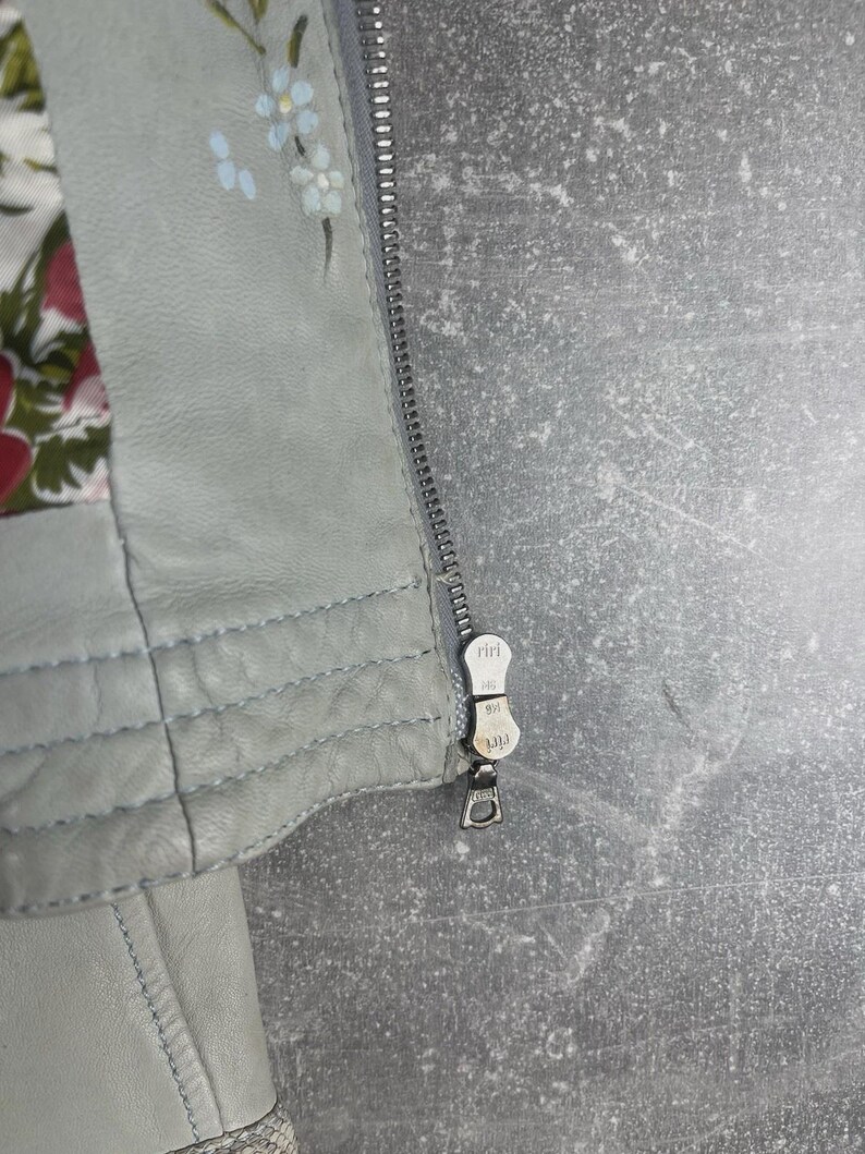 Peut inclure: Gros plan sur une veste en cuir gris clair avec une fermeture &eacute;clair argent&eacute;e. La veste pr&eacute;sente des d&eacute;tails floraux peints &agrave; la main en bleu et blanc. La doublure int&eacute;rieure affiche un motif floral rouge, rose et vert.