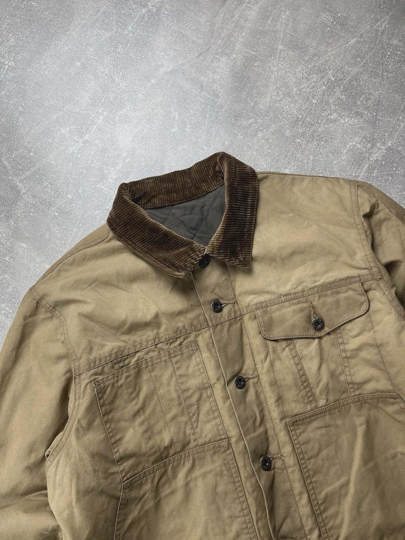 Puede incluir: Una chaqueta de color tostado con cuello de pana marr&oacute;n oscuro, cierre frontal de botones y dos bolsillos en el pecho con solapas abotonadas. La prenda tiene un forro interior acolchado visible en el cuello y costuras detalladas.