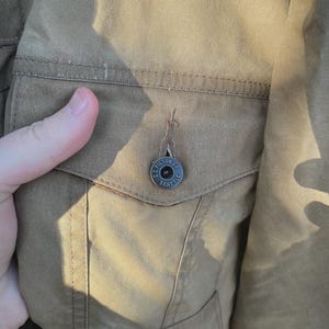 Puede incluir: Una chaqueta de color tostado que presenta un bolsillo en el pecho con cierre de solapa. El bolsillo est&aacute; asegurado por un bot&oacute;n de metal oscuro grabado con el texto C.C. FILSON CO. SEATTLE. La tela tiene un tejido visible y costuras tonales.
