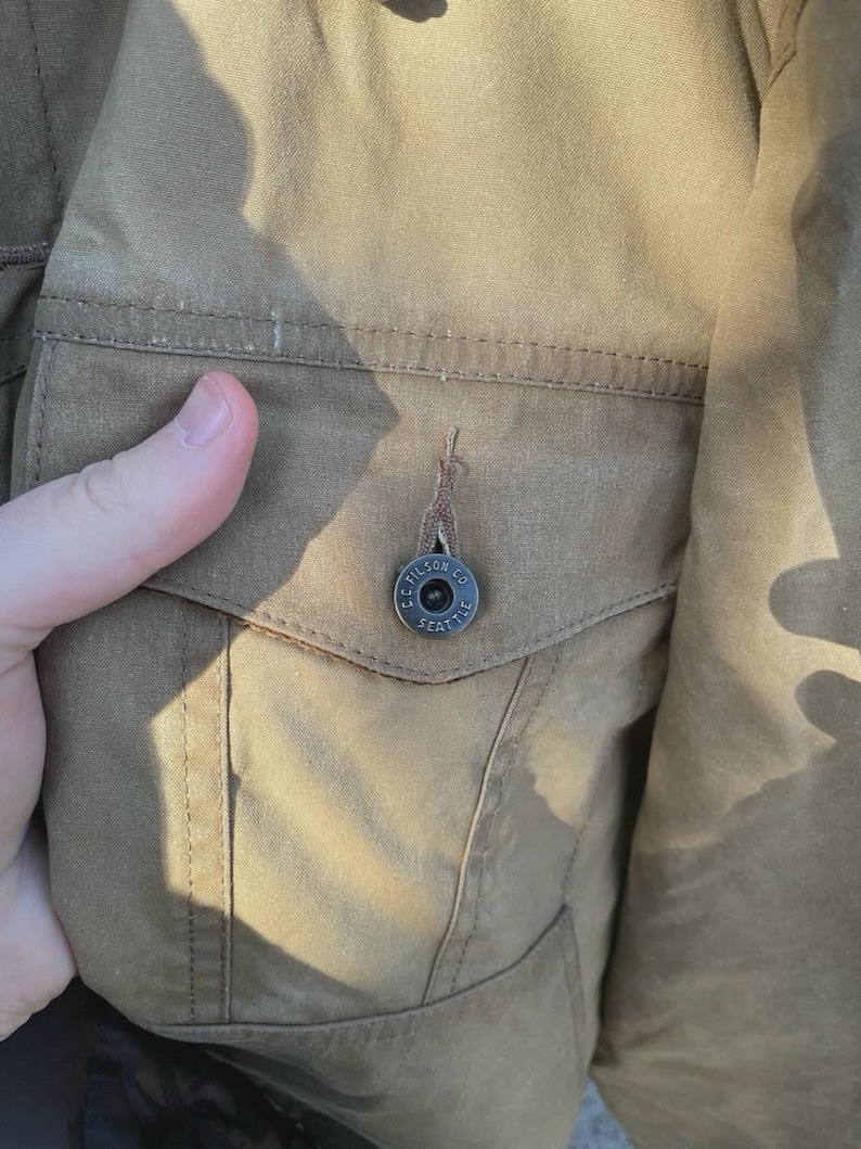Puede incluir: Una chaqueta de color tostado que presenta un bolsillo en el pecho con cierre de solapa. El bolsillo est&aacute; asegurado por un bot&oacute;n de metal oscuro grabado con el texto C.C. FILSON CO. SEATTLE. La tela tiene un tejido visible y costuras tonales.