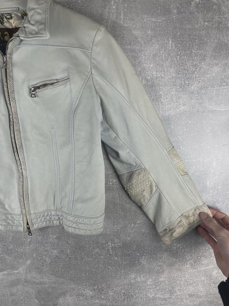 Peut inclure: Veste en cuir gris clair avec fermeture &eacute;clair et petite poche zipp&eacute;e sur la poitrine. La veste pr&eacute;sente des empi&egrave;cements &agrave; motifs peau de serpent sur les coudes et les poignets. La veste est pr&eacute;sent&eacute;e sur fond gris.