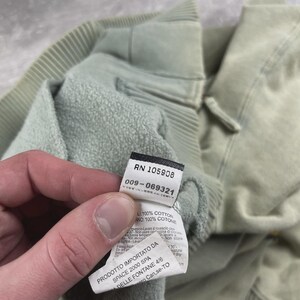 May include: Close-up of a pale green sweatshirt with a label. The label shows the numbers "RN 105908" and "009-069321". The sweatshirt is made of 100% cotton.