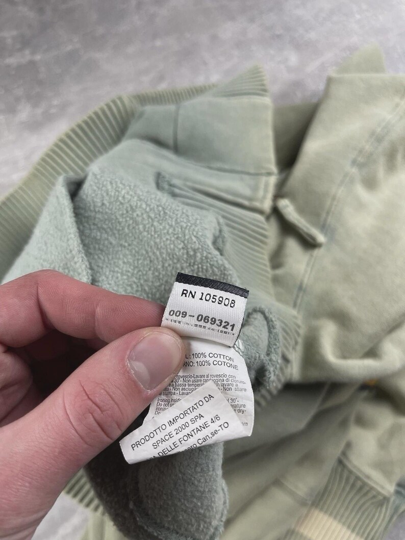 May include: Close-up of a pale green sweatshirt with a label. The label shows the numbers "RN 105908" and "009-069321". The sweatshirt is made of 100% cotton.