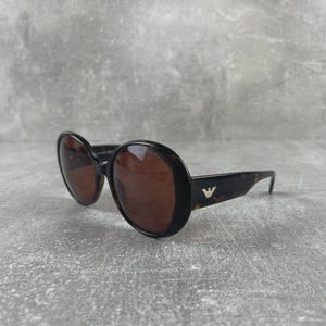 Könnte beinhalten: Runde Sonnenbrille mit braunen Gläsern und einem dunklen Schildpattrahmen. Die Sonnenbrille hat ein kleines silbernes Logo am Bügel. Die Brille ist vor einem grauen Hintergrund platziert.