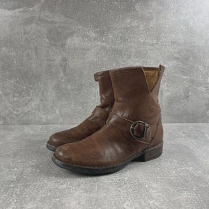 May include: A pair of brown leather ankle boots featuring a rounded toe, a low stacked heel, and a decorative strap across the midfoot secured with a circular metal buckle. The boots have a notched collar design and a dark brown rubber outsole.