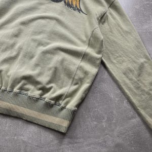 May include: A sage green sweatshirt featuring a graphic of an eye with yellow wings on the back. The bottom hem has a striped design in beige and white. The sweatshirt has long sleeves and a ribbed hem and cuffs.