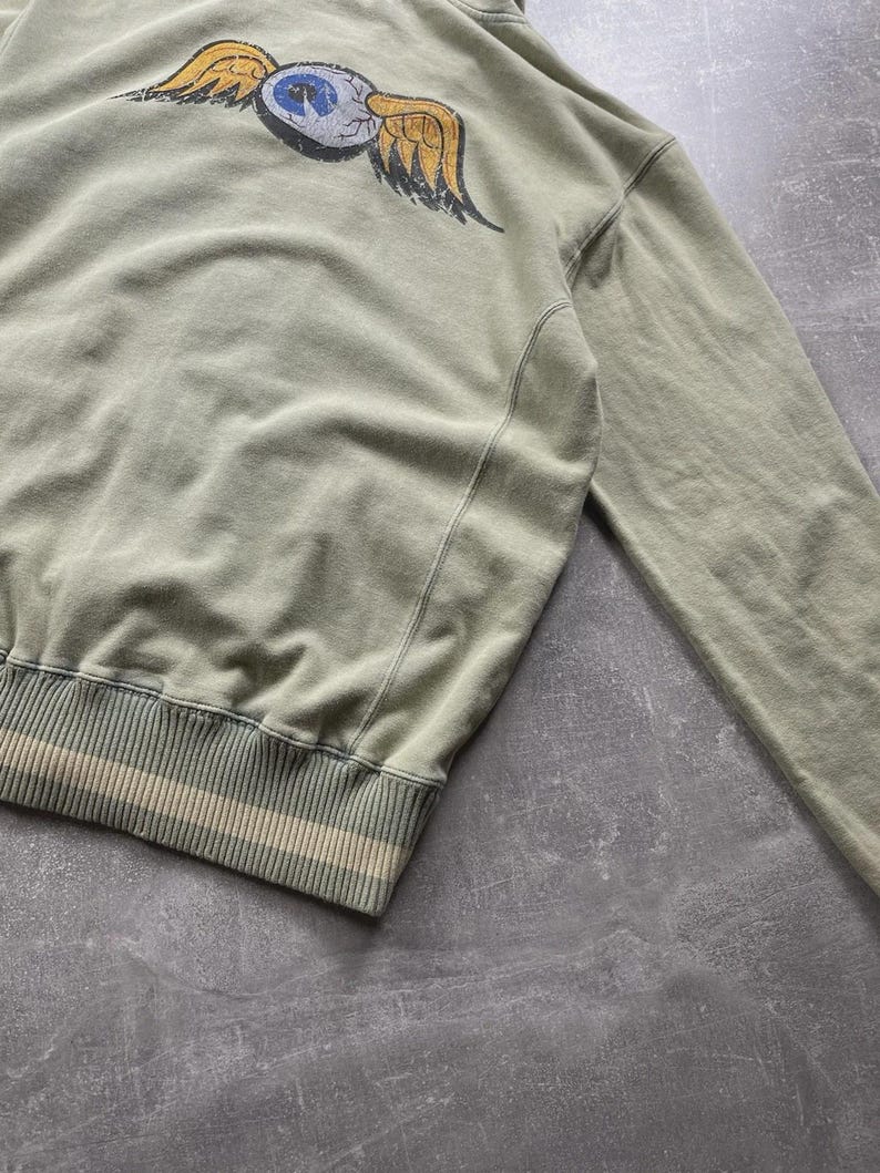 May include: A sage green sweatshirt featuring a graphic of an eye with yellow wings on the back. The bottom hem has a striped design in beige and white. The sweatshirt has long sleeves and a ribbed hem and cuffs.