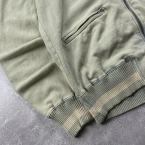 May include: A sage green zip-up jacket with a ribbed cuff and waistband. The jacket features a front pocket with a zip closure. The fabric has a faded, vintage appearance.