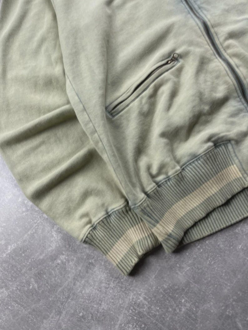 May include: A sage green zip-up jacket with a ribbed cuff and waistband. The jacket features a front pocket with a zip closure. The fabric has a faded, vintage appearance.