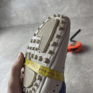 May include: White leather shoe with blue detailing and a studded sole. A yellow measuring tape shows a measurement of approximately 28 cm. The shoe has decorative perforations and a textured sole.
