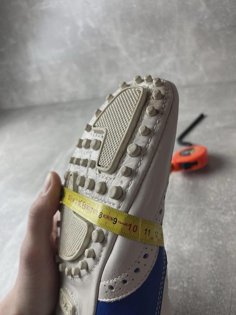 May include: White leather shoe with blue detailing and a studded sole. A yellow measuring tape shows a measurement of approximately 28 cm. The shoe has decorative perforations and a textured sole.