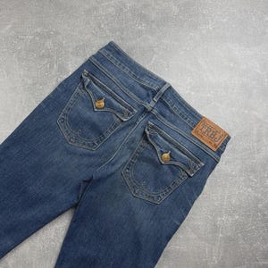 May include: Blue denim jeans featuring two back pockets with button-down flaps, decorative stitching, and a brown leather patch on the waistband embossed with the text TRBJ. The jeans have a classic medium wash finish and visible belt loops.